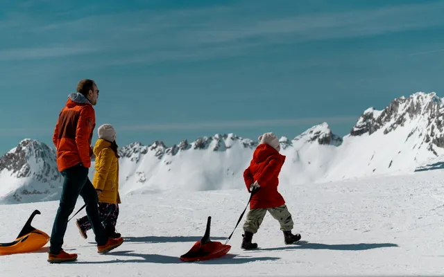 A parent and two children carrying sledges across a sunny snowfield